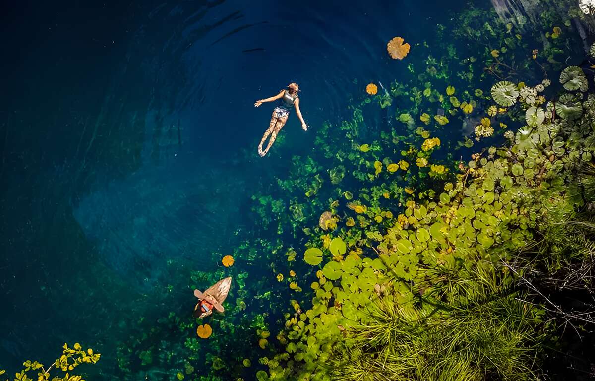 A woman swimming and a woman kayaking in a turquoise blue water cenote. Circular green leaves floating on the right side.