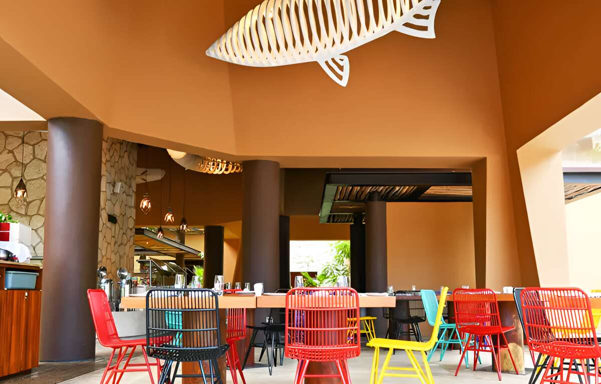A well-lit restaurant. A decorative fish hanging from the ceiling. Colorful chairs around a table.