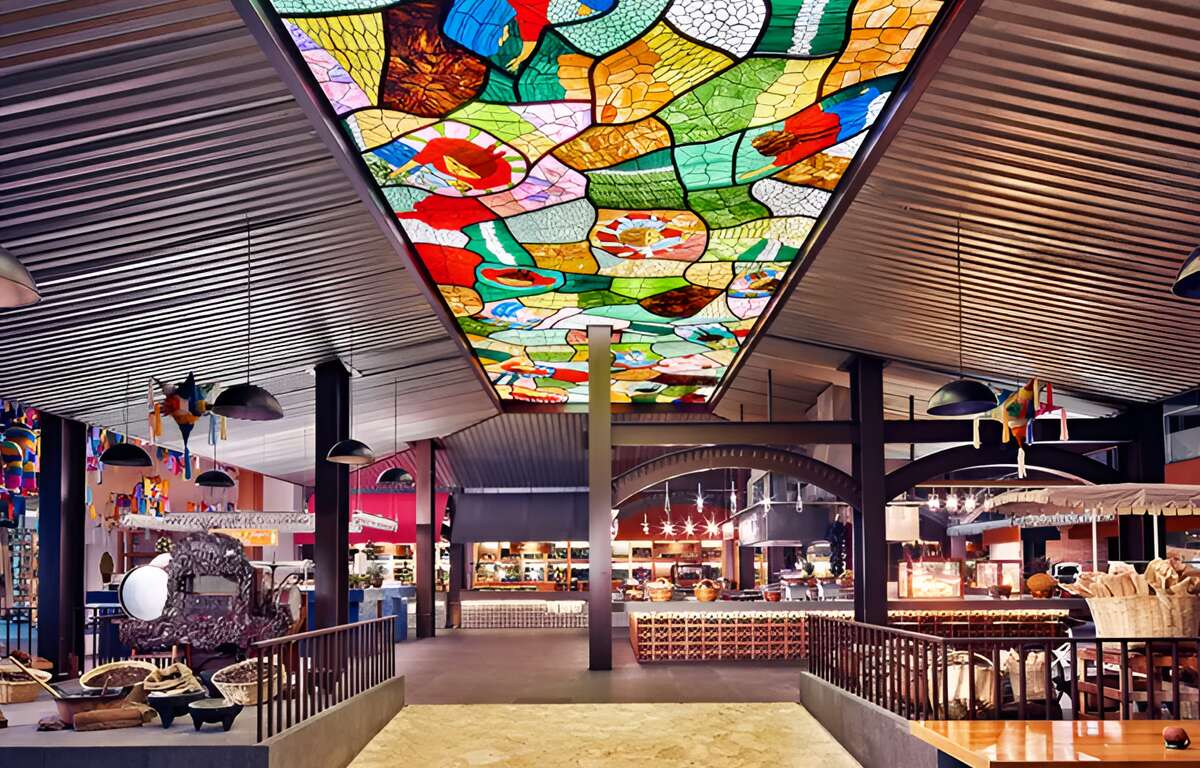A stained glass window covers a large part of a restaurant. Tables and chairs on the sides. In the background several Mexican food stalls.