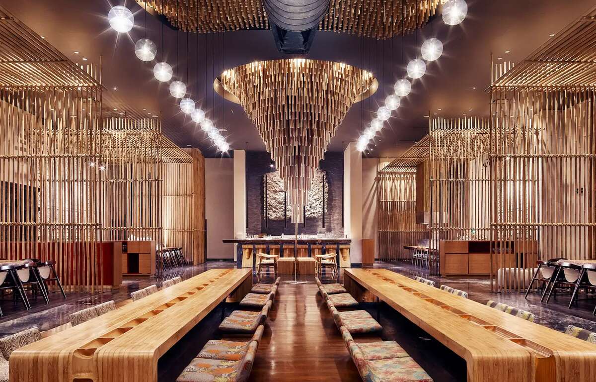 A fancy Japanese style restaurant. Two rows of long wooden tables with chairs on each side. A contemporary circular ornament hangs from the ceiling.