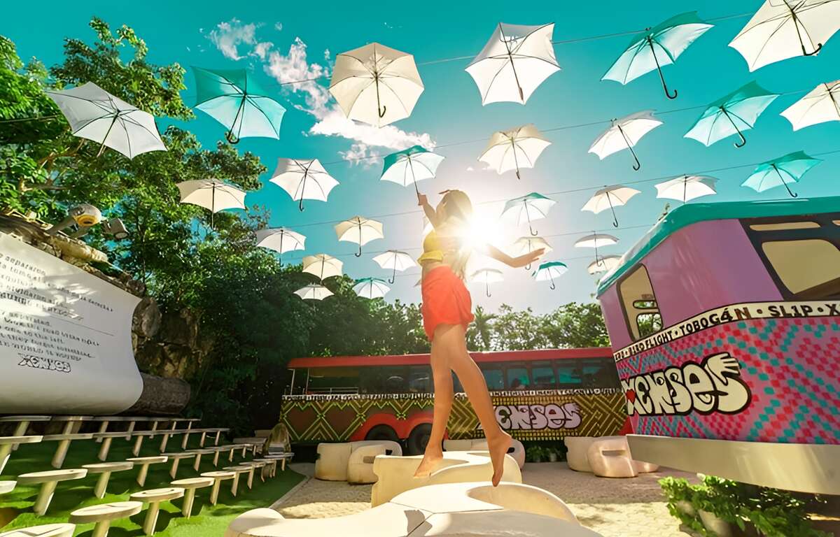 Umbrellas hanging in 5 lines, a woman reaching for one of them. Two buses with the Xenses logo, one in the background and one on the right side.
